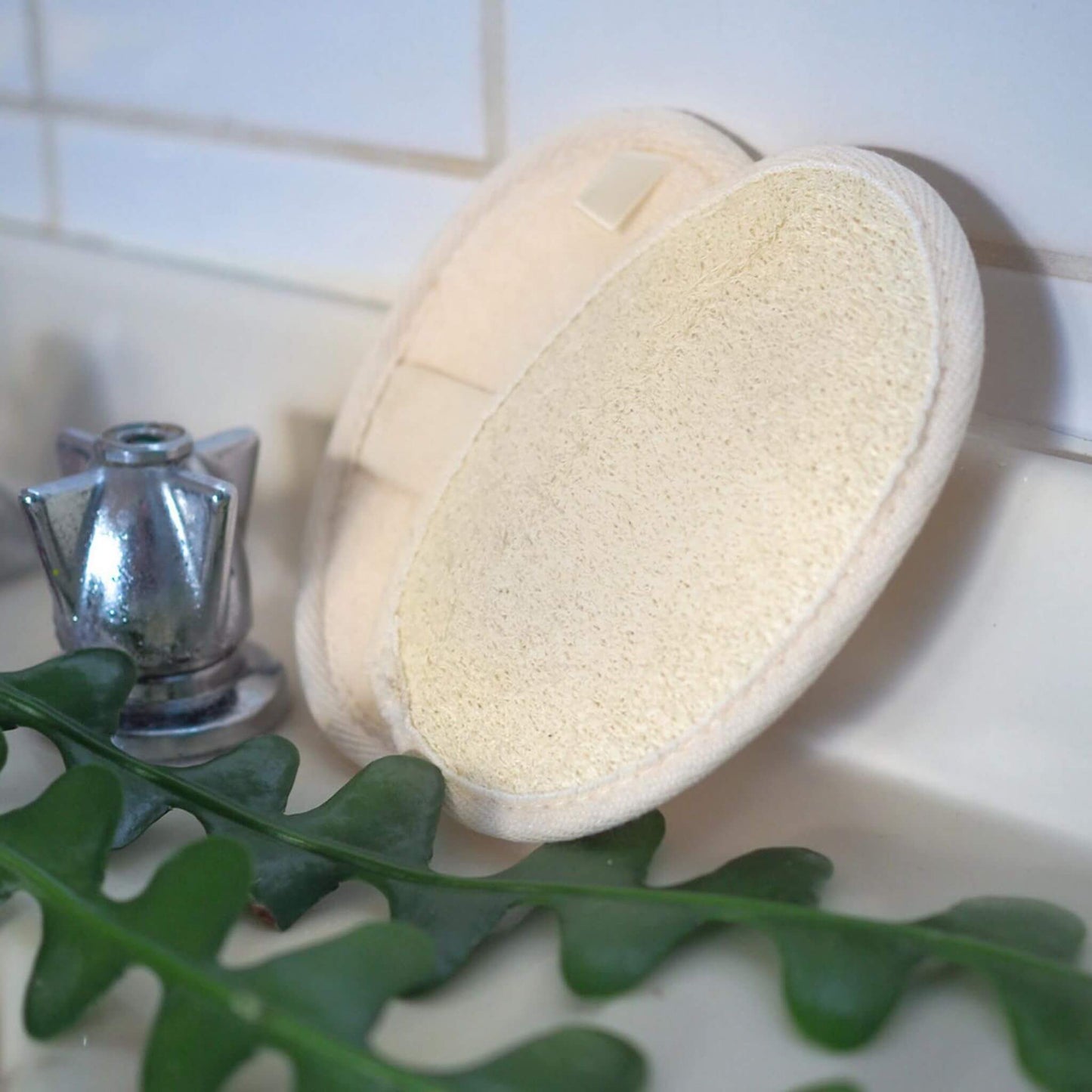 natural loofah body scrub with elastic handle for holding. Sitting on sink with tap and green leaves